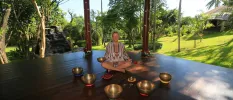 Smiling women with her eyes closed sits among sound bowls cross-legged in an open-air pavilion surrounded by gardens