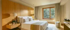 Neutral hotel bedroom with a wood-panelled back wall, white bedding, rattan hanging lights and a window with a view of greenery