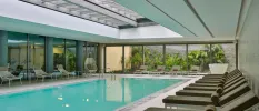 Rectangular indoor pool with floor-to-ceiling windows and grey-cushioned loungers lined up along the right-hand side