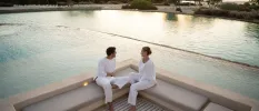 Man and woman in white relax on a neutral sofa next to a pool and beach as the sun sets behind them