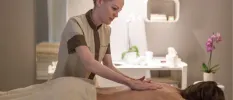 Smiling spa therapist massages a woman's back in a neutral treatment room with a pink potted flower and white vanity unit