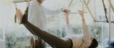 Woman lies on her back with her arms and feet outstretched in a reformer Pilates machine, as an expert instructor guides her