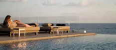 Woman relaxing on a cushioned lounger next to an infinity pool with a glass-walled edge and a sea view