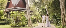 Man and woman stroll through tropical gardens hand in hand, with a pavilion to one side