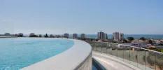 Swimming pool with a curved infinity edge, overlooking a glass balcony and a high-rise seafront town in the distance