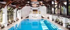 Rectangular indoor pool with a dark wood-beamed ceiling and armchair-style seating lined up at the water's edge