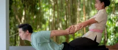 Thai therapist pulls back the arms of a man lying on his front as part of a massage treatment in tropical gardens