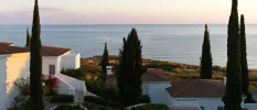 Whitewashed buildings with terracotta roof tiles, surrounded by cypress trees and with a sea view 
