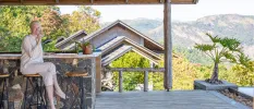 Smiling woman sips from a cup as she sits on an outdoor terrace overlooking green-cloaked mountains