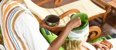 Woman sits in a head wrap consisting of large green leaves, as a therapist drips oil onto her head in a traditional Ayurveda treatment