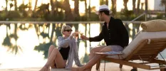 Couple toasting with a drink by the pool as the sun sets beyond the palm trees behind