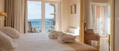 Neutral hotel bedroom with cream walls, curtains and white bedding, a wooden doorway showing an armchair, plus a balcony with ocean views