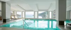 Indoor swimming pool with steps leading into it, with a terrace with two white-cushioned loungers and windows with a view of the sea and hills beyond