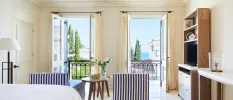 Stylish hotel room with blue and white pinstriped armchairs, a wooden bookcase with a TV, and a small wooden table overlooking a balcony with black wrought-iron railings and a a view of cypress trees and the sea