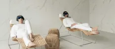 A man and woman in white clothes lying on wooden loungers wearing black eye masks and headphones, in a room with white marble walls