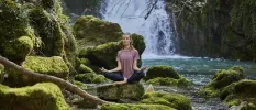 Woman in dark leggings and a purple T shirt meditating on a rock surrounded by other rocks and a waterfall