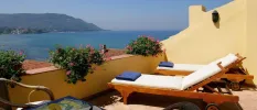 Sunny terrace with wooden white-cushioned loungers and blue towels, overlooking the sea and mountains 