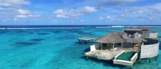 Thatched overwater villa in turquoise shallows, with a wooden deck with green cushions and a swimming pool
