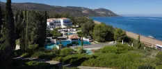 Cypress trees and lush shrubs cloaking a whitewashed resort and central swimming pool, with a gold sandy beach and blue sea in the background