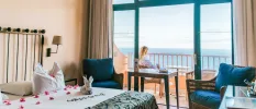 Hotel room with cream walls, white bedding with pink rose petals, a blue armchair and a balcony where a woman sits looking out to sea