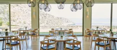 Airy restaurant with floor-to-ceiling windows with views of the sea and craggy cliffs, simple wooden tables and chairs and a cluster of contemporary ceiling pendant lights in a wiry black design