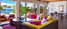 Villa interior with a dark-wood sofa with yellow and pink cushions, a glass coffee table with a driftwood-design bottom, a dining table behind, wood-beamed ceiling and full-length windows overlooking the beach and ocean