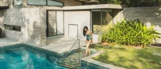 Woman with long dark hair in a dark swimsuit steps into a pool outside a contemporary villa