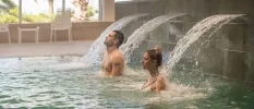 Man and woman relax under water jets spouting out of a tiled rectangular wall in an indoor pool
