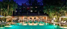 Night-time at an outdoor pool, surrounded by white loungers and umbrellas and tropical greenery, in front of a al fresco restaurant terrace