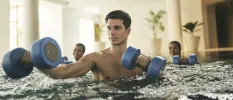 Man exercises with blue dumbbells in an indoor swimming pool with two people behind him