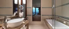 Neutral tiled bathroom with marble finishes, a rectangular bathtub, vanity unit with a large white sink, glass bottled amenities and a vase of orchids