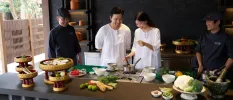 Two guests in white experiment at a cooking station as two chefs look on
