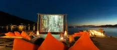 Large cinema screen in front of wooden loungers with orange cushions, next to a wooden jetty on the seafront under a night sky