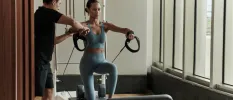 Woman in grey leggings and a crop top does resistance training as a trainer helps to extend