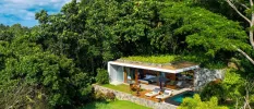 Villa covered in and surrounded by greenery, with a bed overlooking the view and a wooden terrace with cushioned loungers and a pool