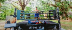Muay Thai boxer stands in a ring in blue shorts in a leafy garden, with a sign next to him detailing 'private sessions on request'