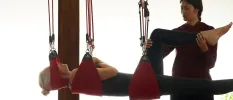 Woman in active wear lies in a red harness-like system as a therapist bends her leg in the air
