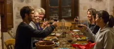 Smiling group clink glasses of red wine in a stone-walled room over a table filled with colourful food