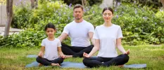 Man, woman and little girl in white t shirts and black trousers meditate on a lawn in a garden