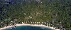 Green-cloaked hillside with a white sandy beach and sea at the bottom, with rows of thatched-roof villas lined up towards the shoreline