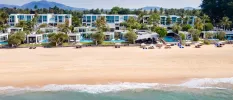 Low-rise white buildings lined up along a beach with private pools and floor-to-ceiling windows, surrounded by palm trees