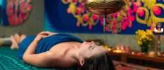 Woman lying down in a blue towel with oil pouring onto her head from a gold jug abovr