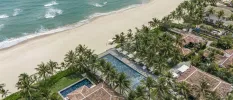 A beachfront resort covered in palm trees overlooking ivory sands, with tile-roofed buildings and swimming pools as seen from above