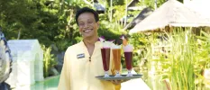Smiling staff member in yellow carries a tray of tall, colourful drinks through tropical grounds