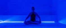 Woman in a black swimsuit sits cross-legged in a blue-lit room