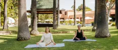Two women sit in yoga poses on blue mats in a tropical garden
