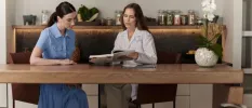 Two women sitting at a wooden table looking at a book, in front of a table full of jars