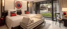 Hotel room with twin beds with white bedding, red and black cushions, dark-wood accents and a balcony door open to pool and garden views