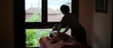 Woman lies on her front with a flower in her hair in a darkened room, as a therapist massages her back