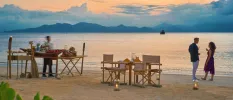 Beachfront at sunset: a couple enjoy a drink by the shoreline with a table set up for two behind them, and a staff member works behind a table full of food